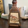 A vintage, textured brown decanter with a round red cap sits on a wooden table. The front features an illustration of a fox in a grassy field. Blurred books and decor are visible in the background.