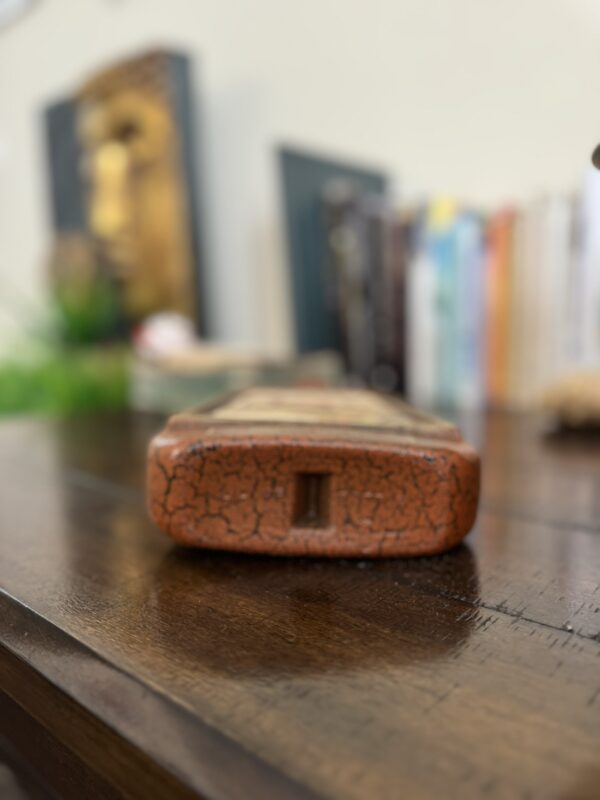 Close-up of a small, rectangular, reddish-brown object with a cracked surface sitting on a wooden table. In the background, various colorful books and blurred decor, including a golden Buddha face, are visible but out of focus.