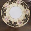 An ornate porcelain plate with a wide black border decorated with gold filigree and clusters of pink, yellow, and blue flowers. The center is plain white with a thin gold rim. The plate rests on a dark wooden surface.