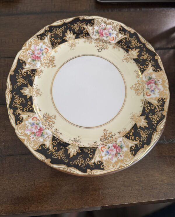 An ornate porcelain plate with a wide black border decorated with gold filigree and clusters of pink, yellow, and blue flowers. The center is plain white with a thin gold rim. The plate rests on a dark wooden surface.