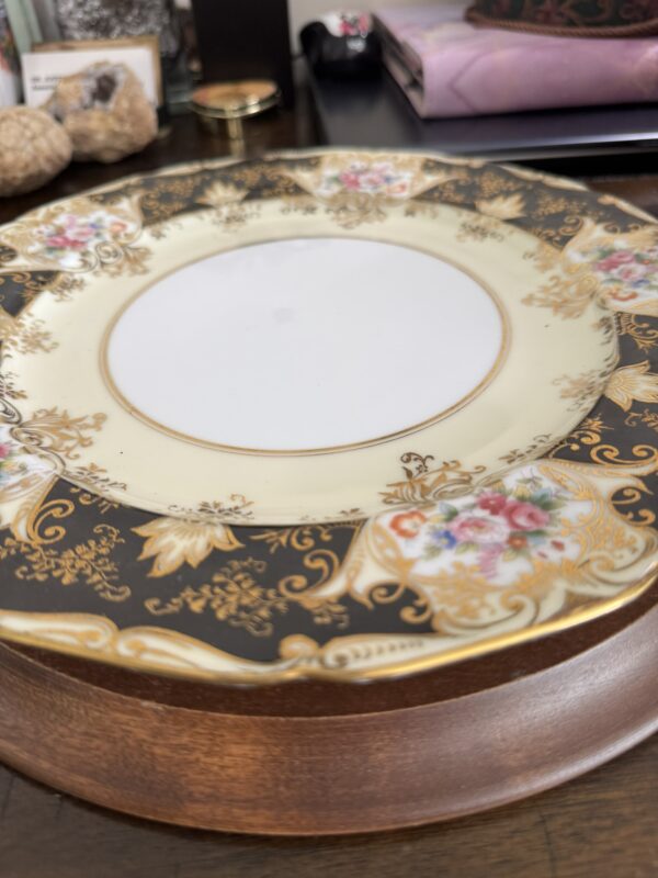 A close-up of an ornate porcelain plate with a gold, black, and cream border featuring floral patterns. The plate sits on a wooden surface, and blurred objects like walnuts, a tin, and books are visible in the background.