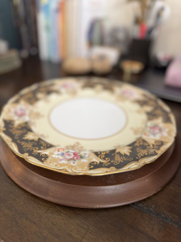 A decorative porcelain plate with a gold and floral border rests on a round wooden tray atop a dark wooden table. The background is softly blurred, showing assorted desk items and books.