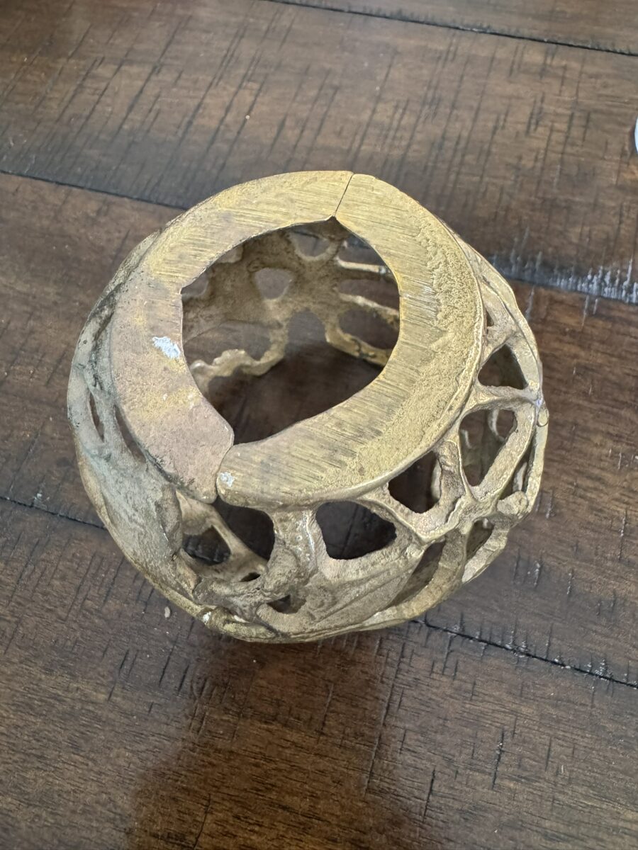 A carved, hollow, spherical wooden object with geometric cutouts sits on a brown wooden table. The top of the sphere has an irregular, open section, and the wood appears weathered with visible texture and small marks.