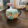 A small, colorful cup with a black handle sits on a wooden table. The cup features painted orange flowers and a bird on a green background. In the background are books, a decorative dish, rocks, and other desk items, all slightly out of focus.