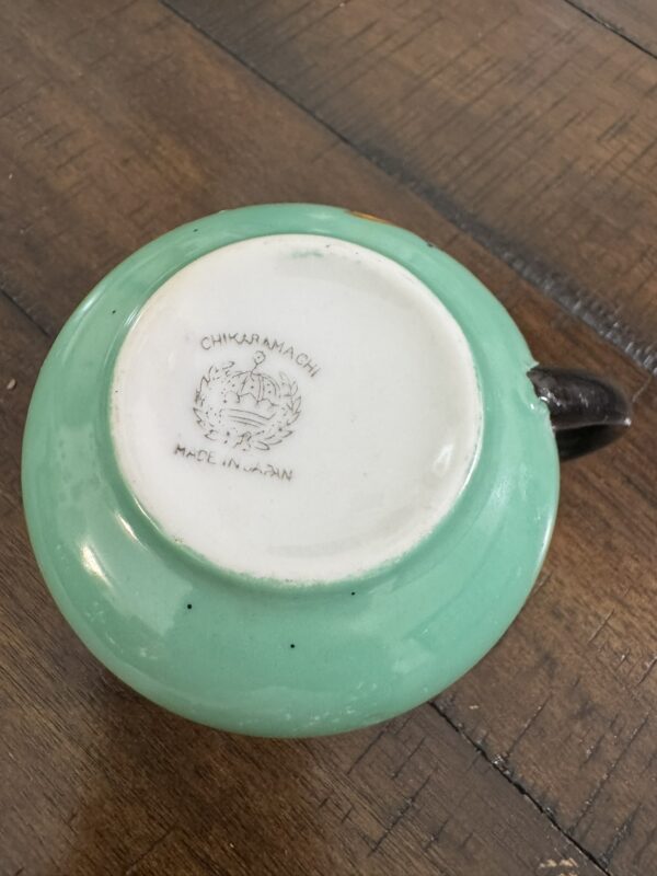 A green ceramic cup lies upside down on a wooden surface, showing its white base. The base is stamped with CHIKARAMACHI MADE IN JAPAN and an image of a phoenix-like bird with outstretched wings. The cup has a black handle.