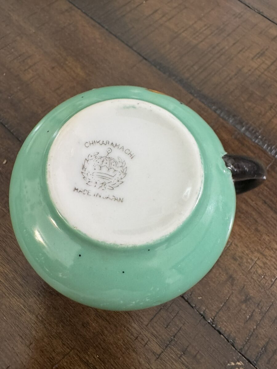 A green ceramic cup lies upside down on a wooden surface, showing its white base. The base is stamped with CHIKARAMACHI MADE IN JAPAN and an image of a phoenix-like bird with outstretched wings. The cup has a black handle.
