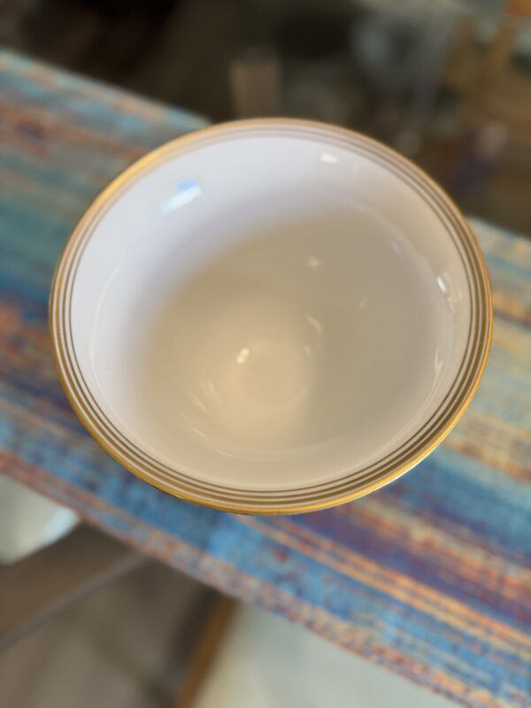 A white ceramic bowl with three gold stripes around the rim sits on a colorful, woven blue table runner. The background is softly blurred, highlighting the bowl as the main focus of the image.