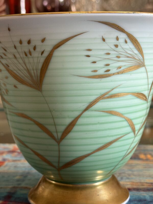 A close-up of a pale green porcelain bowl with delicate horizontal lines and golden botanical designs featuring leaves and seed heads, resting on a gold-colored base. The background is softly blurred, highlighting the bowls intricate detail.
