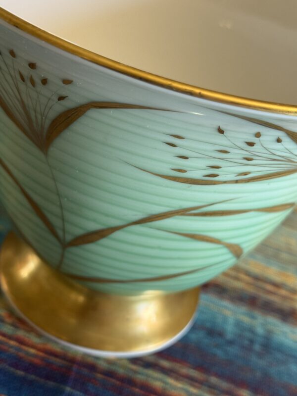 A close-up of a ceramic bowl with a pale green exterior, decorated with delicate gold lines and floral patterns. The rim and foot of the bowl are also gold. The bowl rests on a multicolored, textured fabric surface.