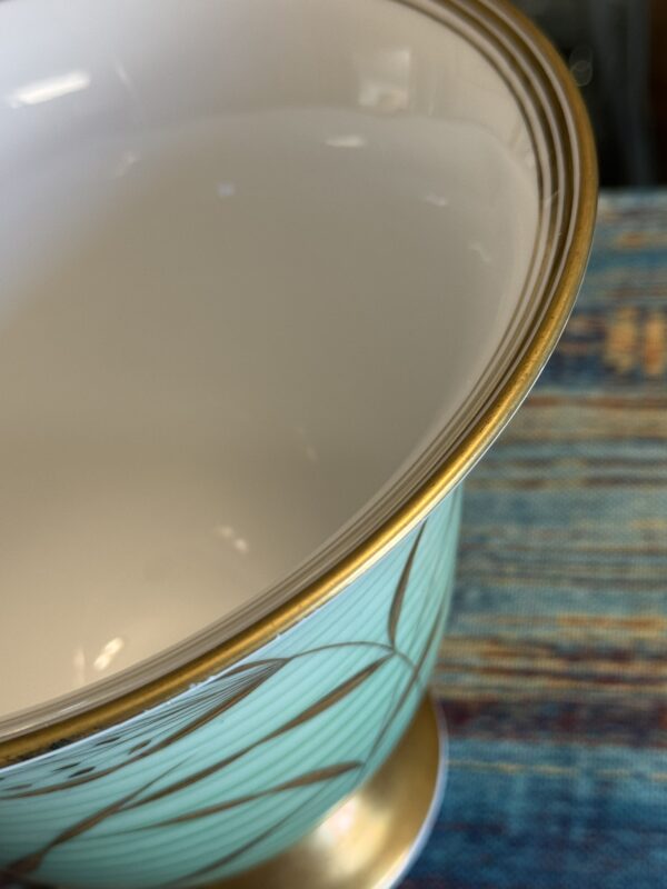 Close-up of a ceramic bowl with a glossy white interior, gold rim, and gold base. The exterior features a light turquoise color with a geometric gold and black linear pattern. The bowl is placed on a woven, multicolored textured surface.