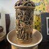 A detailed carved wooden container with an openwork lid displays a scene of a water buffalo amid bamboo and foliage. The container sits in a beige shallow bowl atop a wooden stand, with colorful and monochrome art in the background.