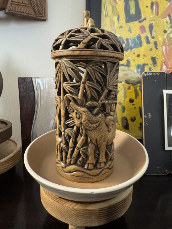 A detailed carved wooden container with an openwork lid displays a scene of a water buffalo amid bamboo and foliage. The container sits in a beige shallow bowl atop a wooden stand, with colorful and monochrome art in the background.