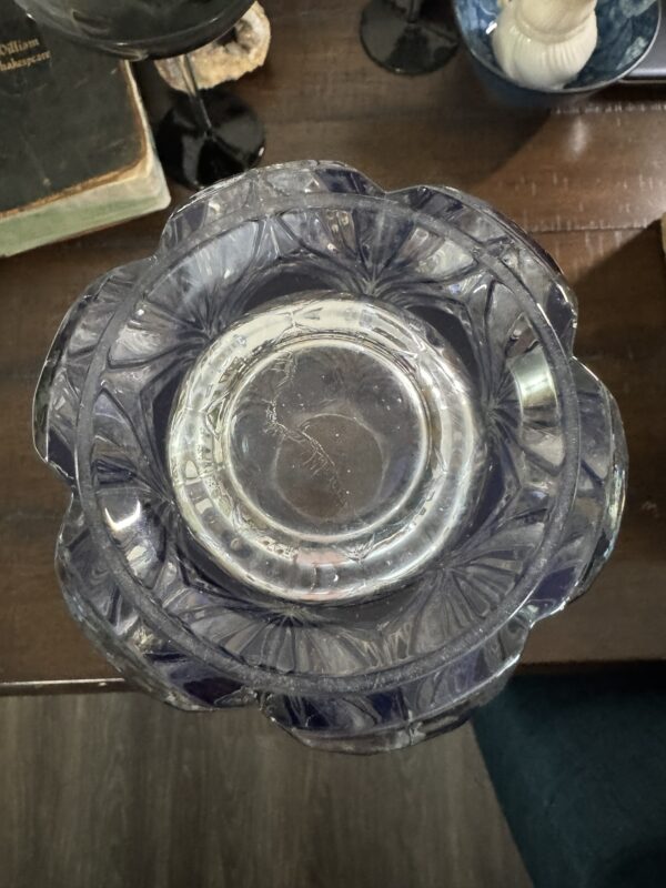 A top-down view of a clear, scalloped-edged glass bowl with intricate patterns, resting on a wooden table. Nearby are a closed book, a black wine glass, a blue dish with a candle, and part of a green chair visible in the lower corner.
