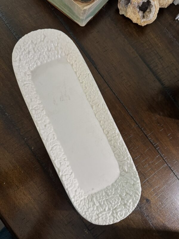 A white rectangular ceramic platter with rounded corners and an ornate, textured floral border sits on a dark wooden table. The platter is empty, and part of a geode and a closed book are visible in the background.
