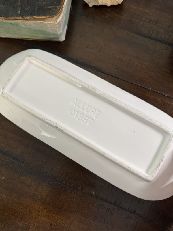 A white ceramic object is shown upside down on a wooden surface. Its base is rectangular and stamped with the text ALLURE © 1997. Part of an old book and a small rock are partially visible nearby.