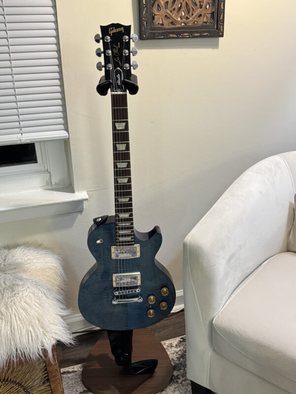 A blue Gibson Les Paul electric guitar on a stand next to a white armchair and window with closed blinds in a cozy, well-lit room.