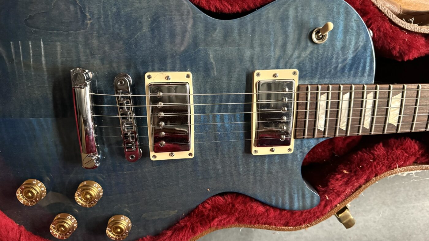 A close-up of a blue electric guitar with gold knobs, chrome hardware, and cream pickup frames, resting inside a red-lined hard case.