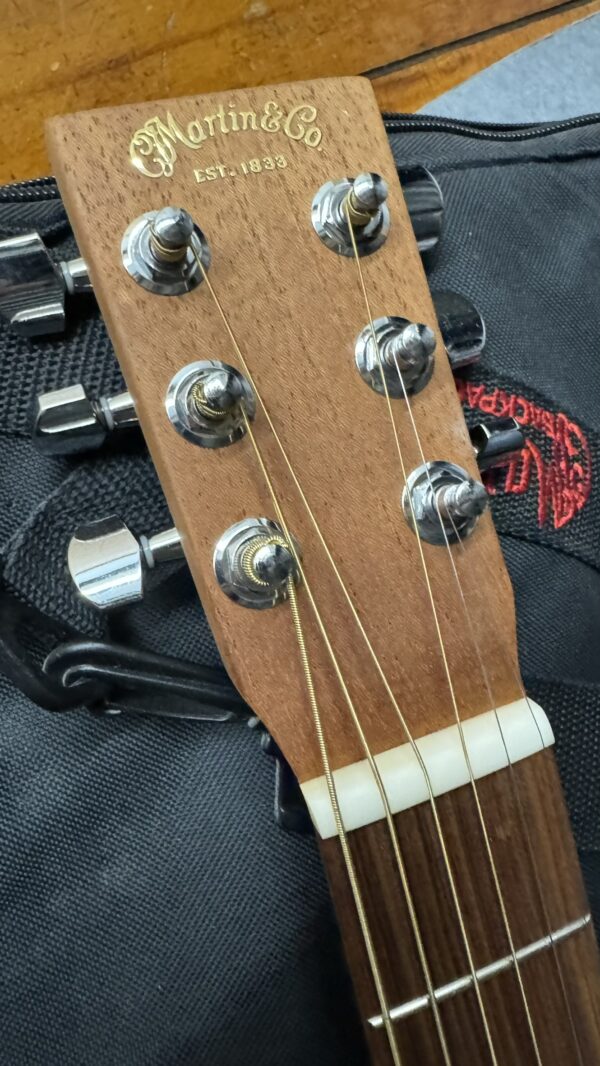 Close-up of a Martin & Co acoustic guitar headstock with tuning pegs and strings, resting on a black fabric bag with visible red stitching.