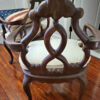 An antique wooden armchair with ornate carvings, a cream seat cushion, and curved legs sits on a hardwood floor. A bench with an American flag cushion is partially visible nearby. White doors are in the background.