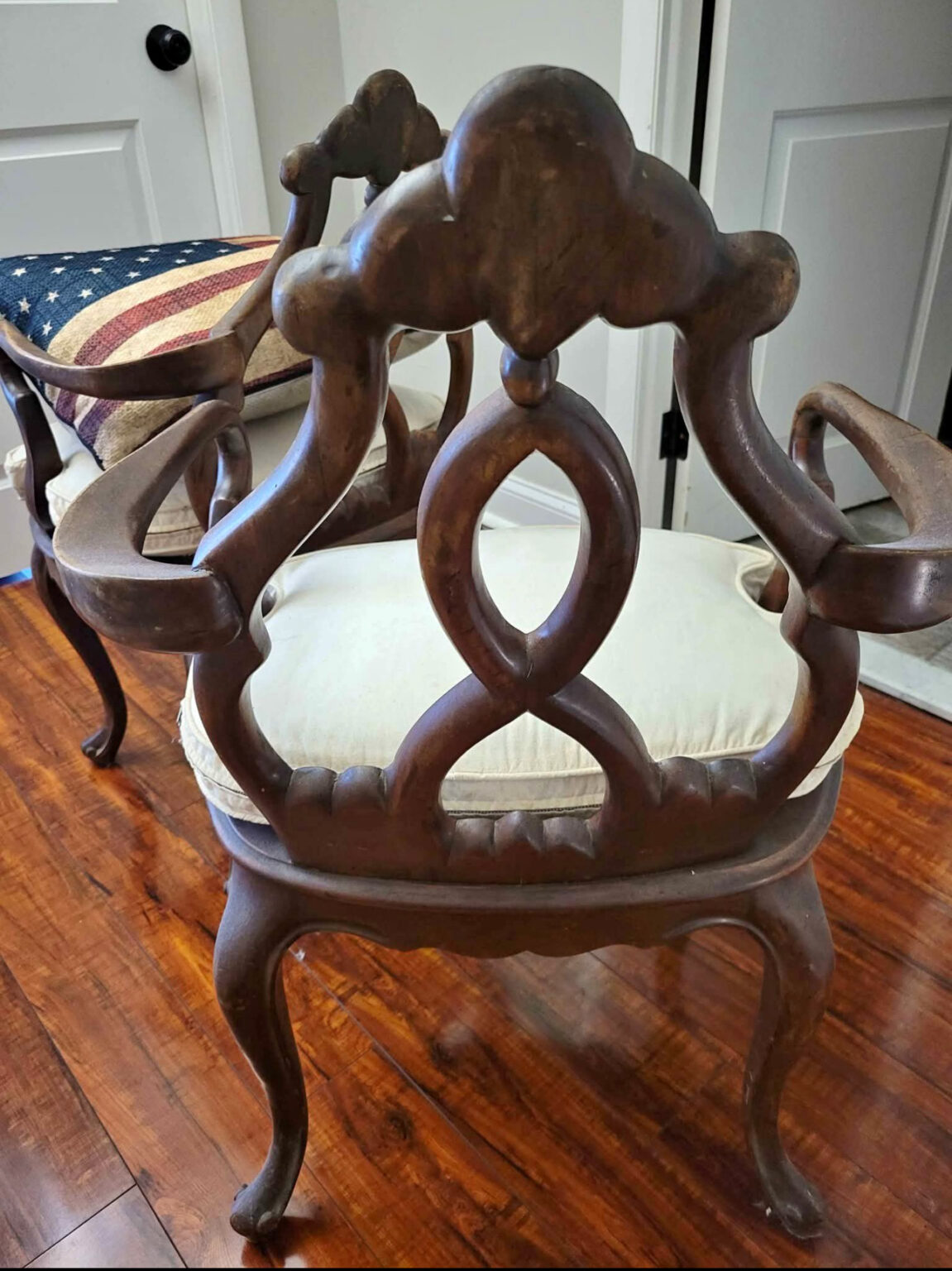 An antique wooden armchair with ornate carvings, a cream seat cushion, and curved legs sits on a hardwood floor. A bench with an American flag cushion is partially visible nearby. White doors are in the background.
