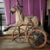 A vintage toy horse with a saddle is mounted on three large wooden wheels, set in front of a purple storage bin and cardboard boxes. The horse appears to be made of wood and metal.
