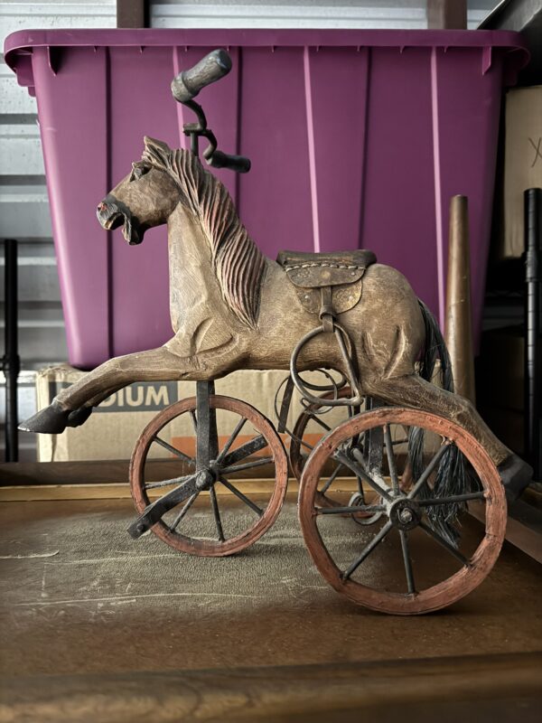 A vintage toy horse with a saddle is mounted on three large wooden wheels, set in front of a purple storage bin and cardboard boxes. The horse appears to be made of wood and metal.