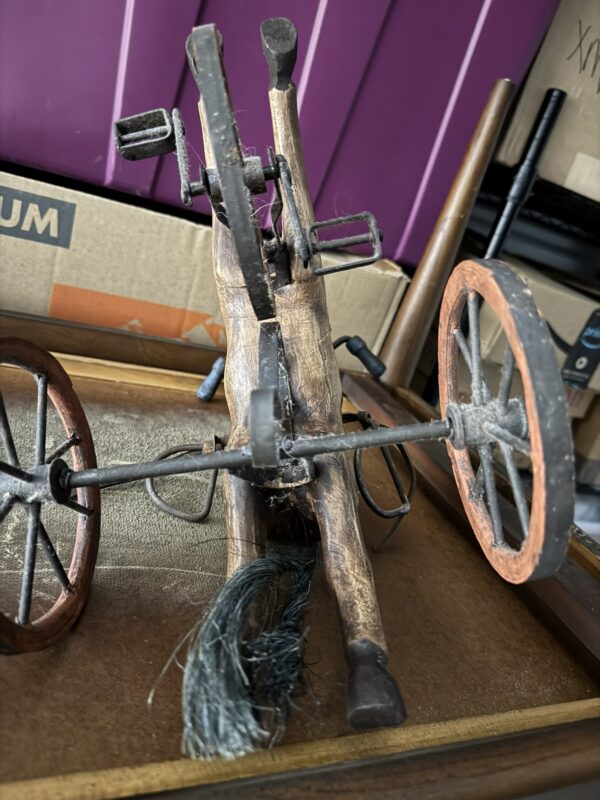A close-up of a rustic, miniature wooden cart with large spoked wheels and metal parts, placed on a surface in front of a cardboard box and a purple striped background.