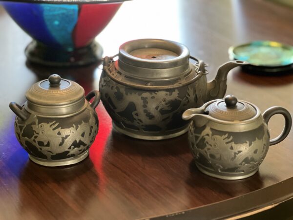 Three ornate, dark metal tea set pieces—including a teapot, sugar bowl, and creamer—feature dragon designs and sit on a polished wooden table, with a colorful bowl and dish blurred in the background.