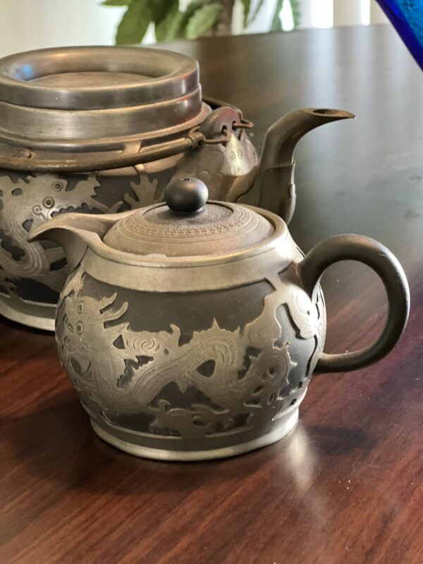 A close-up of an ornate metal teapot with an engraved dragon design, sitting on a wooden table next to a matching teapot. A green plant is blurred in the background.