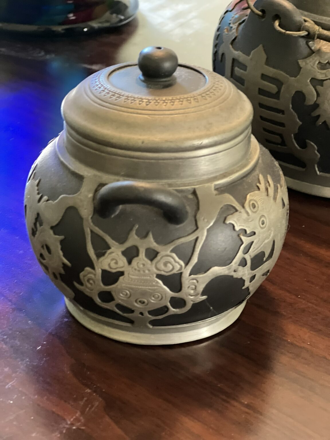 A round, ornately decorated metal jar with a lid, featuring intricate engraved designs and dragon motifs, sits on a dark wooden surface. Another similarly styled container is partially visible in the background.