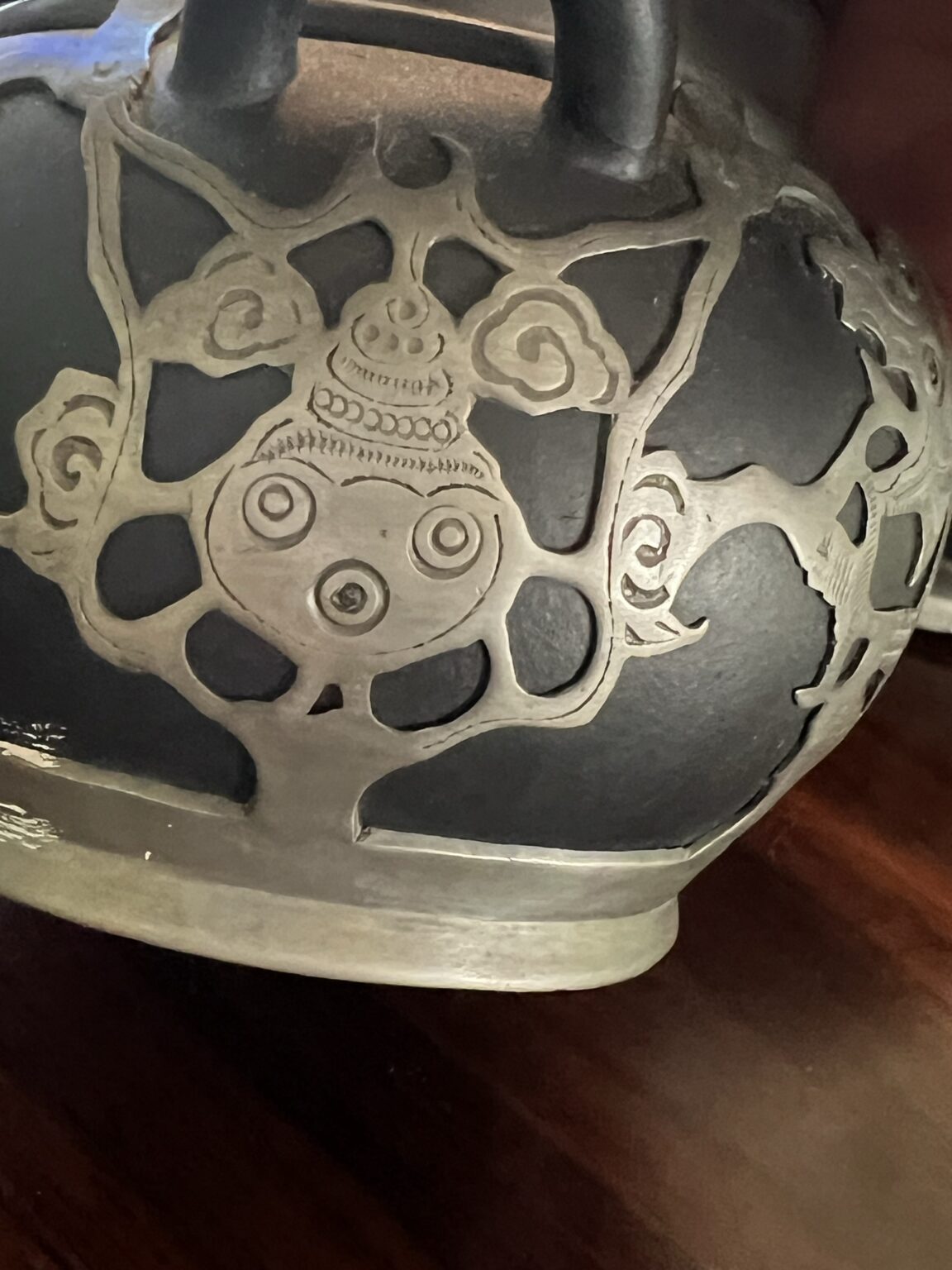 Close-up of a dark metal object with an engraved abstract design featuring swirling shapes, circles, and curved lines, possibly part of a decorative vessel or artifact, placed on a wooden surface.