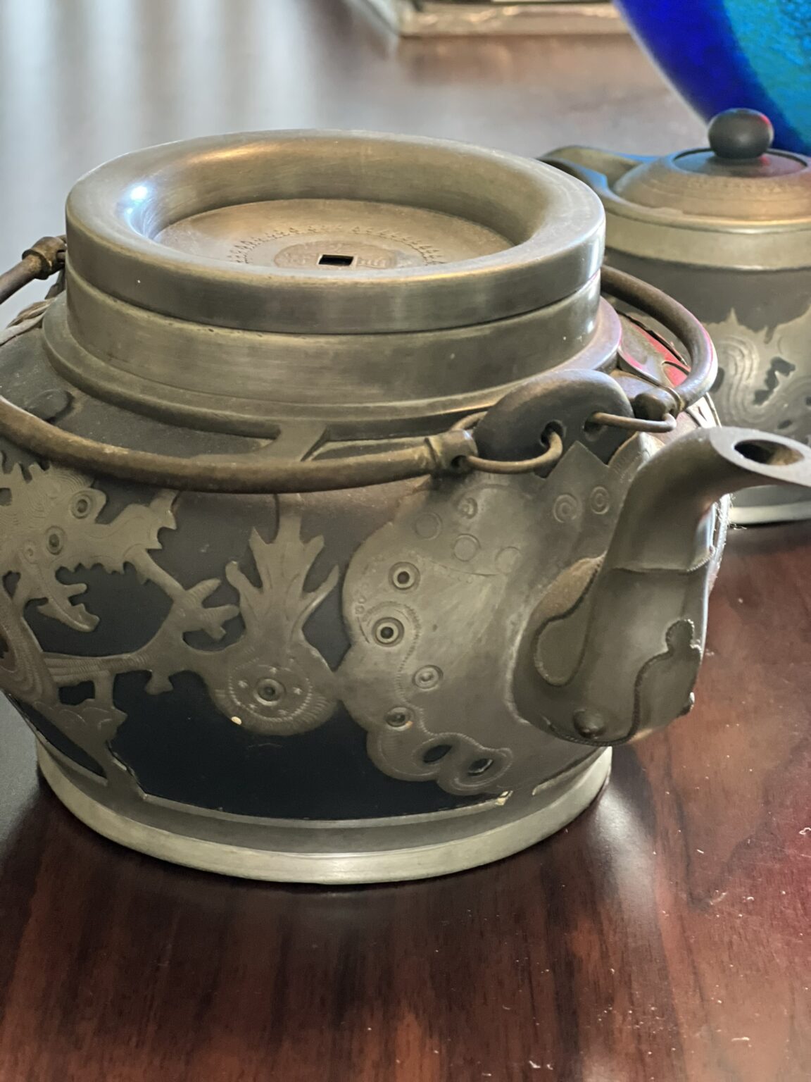 A close-up of an ornate metal teapot with an intricate dragon design on its side, sitting on a wooden table. Another smaller teapot is blurred in the background.