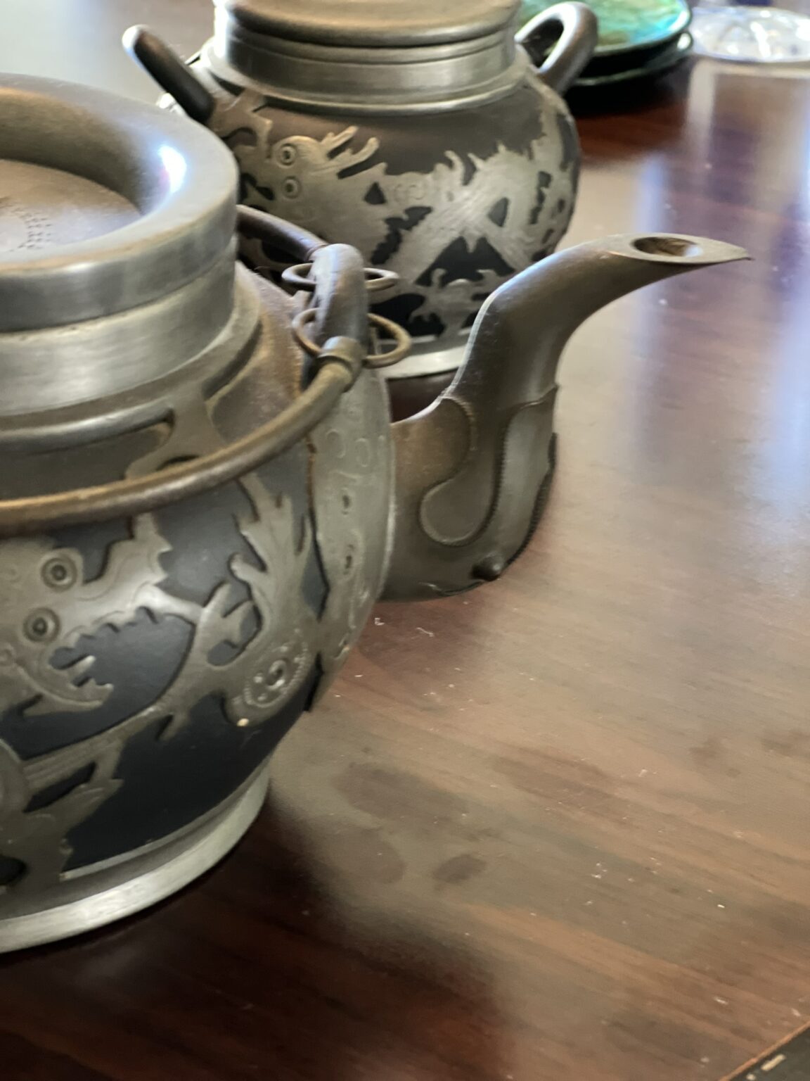 Two dark metal teapots with embossed dragon designs sit on a wooden table. One teapot is in the foreground with its lid visible; the other is slightly behind, showing its curved spout.