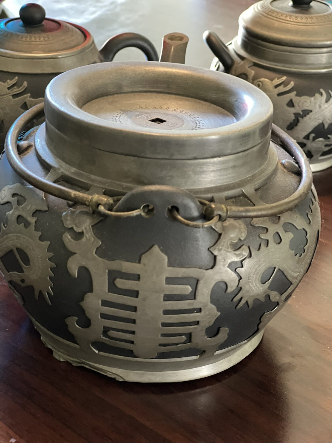 Three ornate metal teapots with intricate designs rest on a dark wooden surface. The closest teapot features a prominent carved pattern and a sturdy handle, while the others are slightly blurred in the background.
