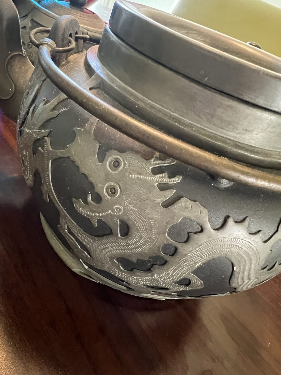 A close-up of a metal pot with a lid, featuring an engraved dragon design on its side, sitting on a dark wooden surface.