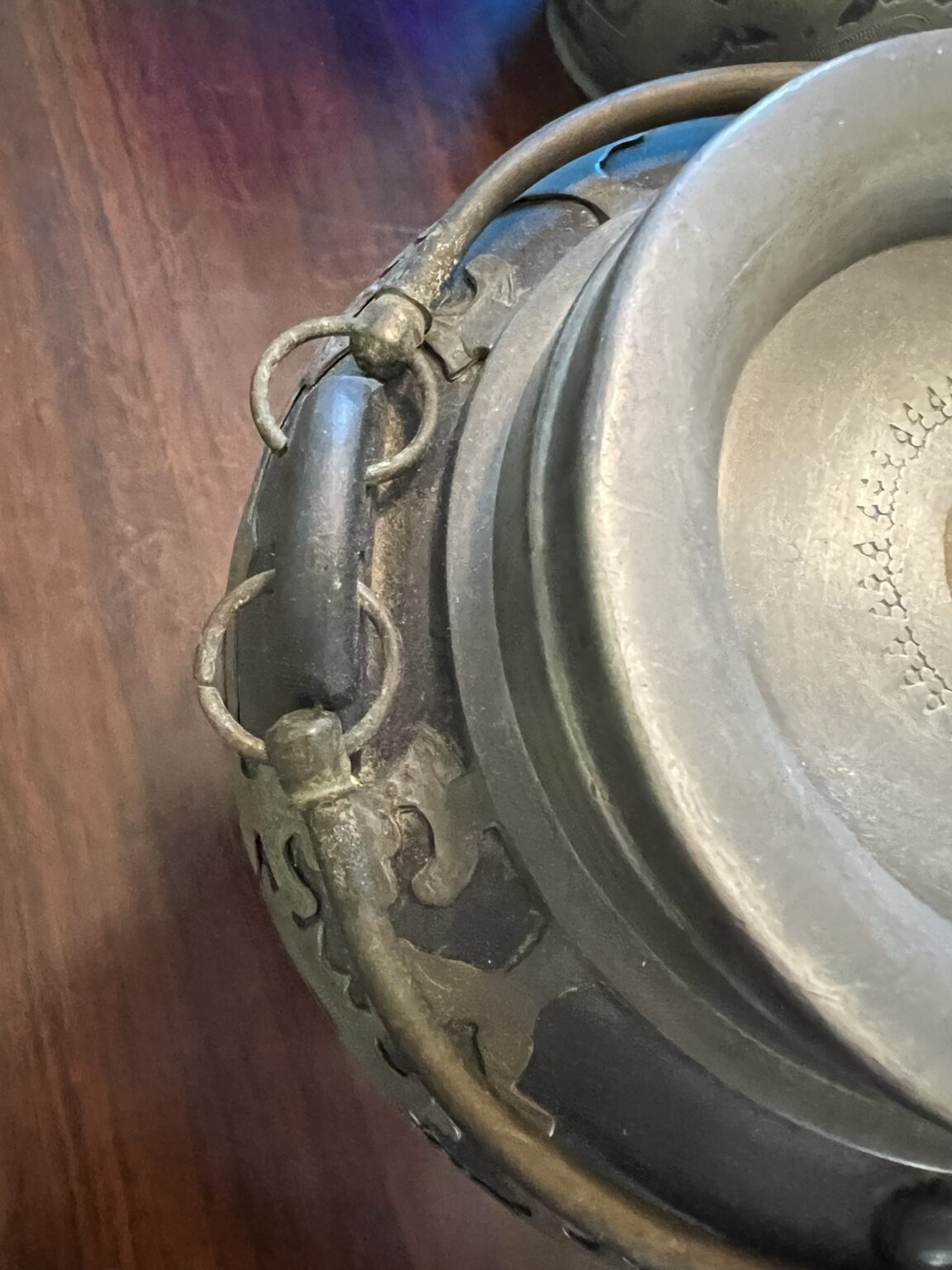 Close-up of a dark metal pot with an ornate handle, attached to the rim by metal rings, resting on a wooden surface. The pot has decorative detailing and appears to be antique or handcrafted.