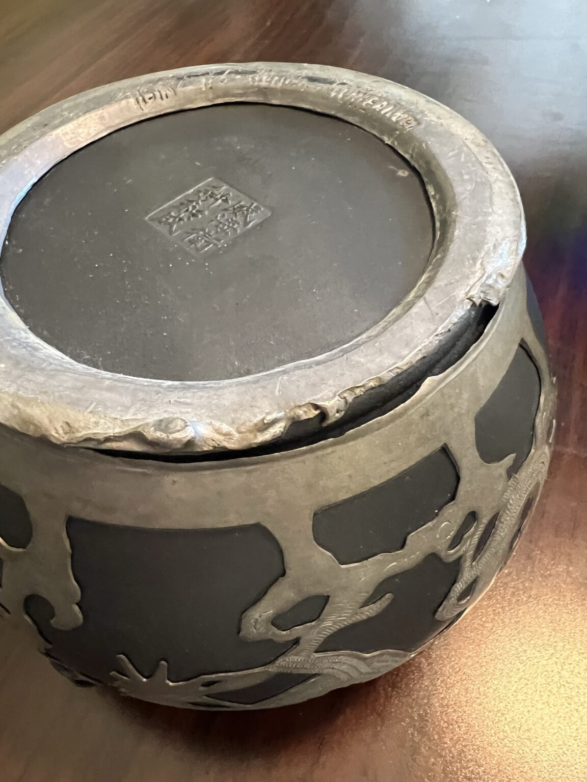 A close-up of the bottom of a black pot with silver metallic decorative accents and a square maker’s mark on the base, sitting on a wooden surface. The upper rim shows signs of wear and rough texture.