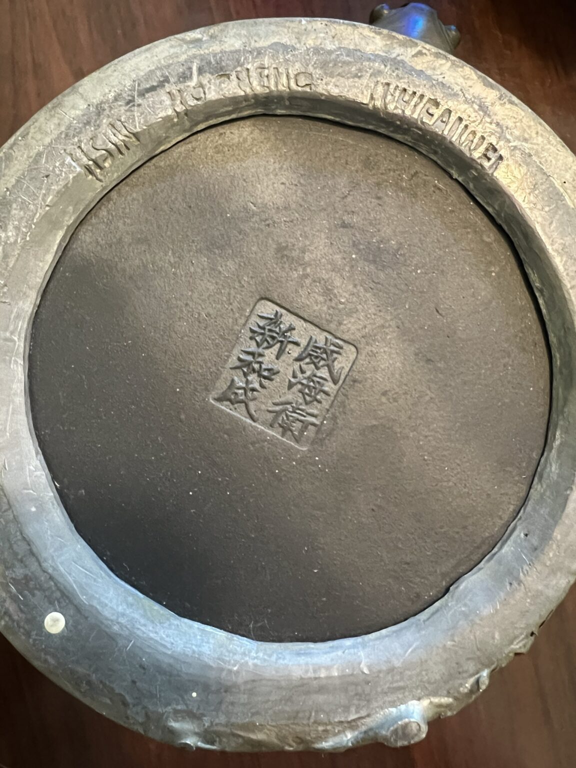 The image shows the bottom of a round, dark ceramic object with a square stamp of Chinese characters in the center and raised English text reading ASIAN IMPORTING WHOLESALERS around the rim.