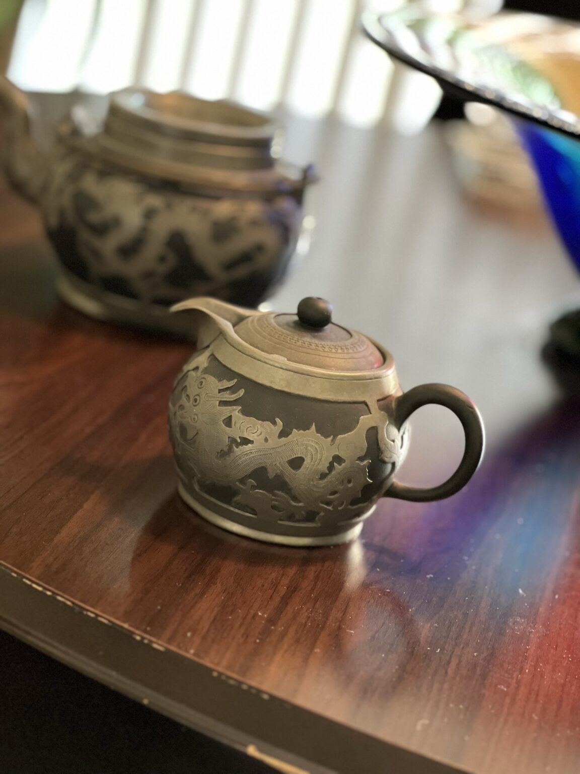 A small, ornate teapot with an engraved dragon design sits on a polished wooden table, with a matching larger teapot blurred in the background.