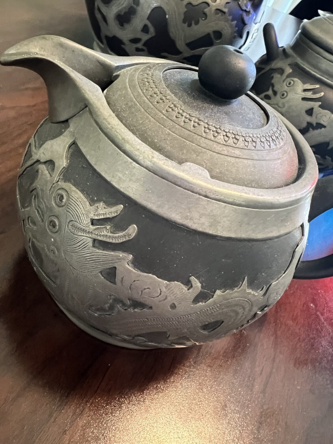 A close-up of a dark, round teapot with a lid and handle, decorated with intricate dragon carvings. The teapot sits on a wooden surface, with another similarly designed teapot partially visible in the background.
