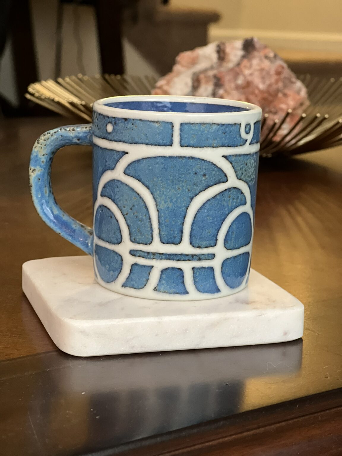 A blue ceramic mug with white geometric patterns sits on a square marble coaster atop a wooden table. A decorative rock and a golden tray are blurred in the background.