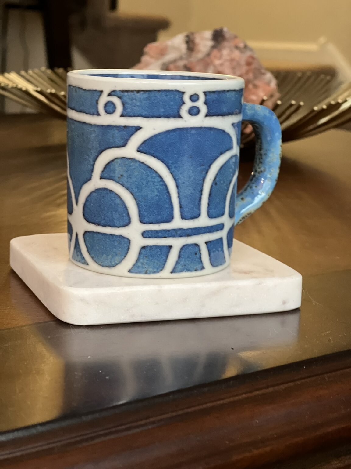A blue mug with a white geometric pattern sits on a square white coaster atop a wooden table. In the background, there is a decorative pink stone and a gold tray.