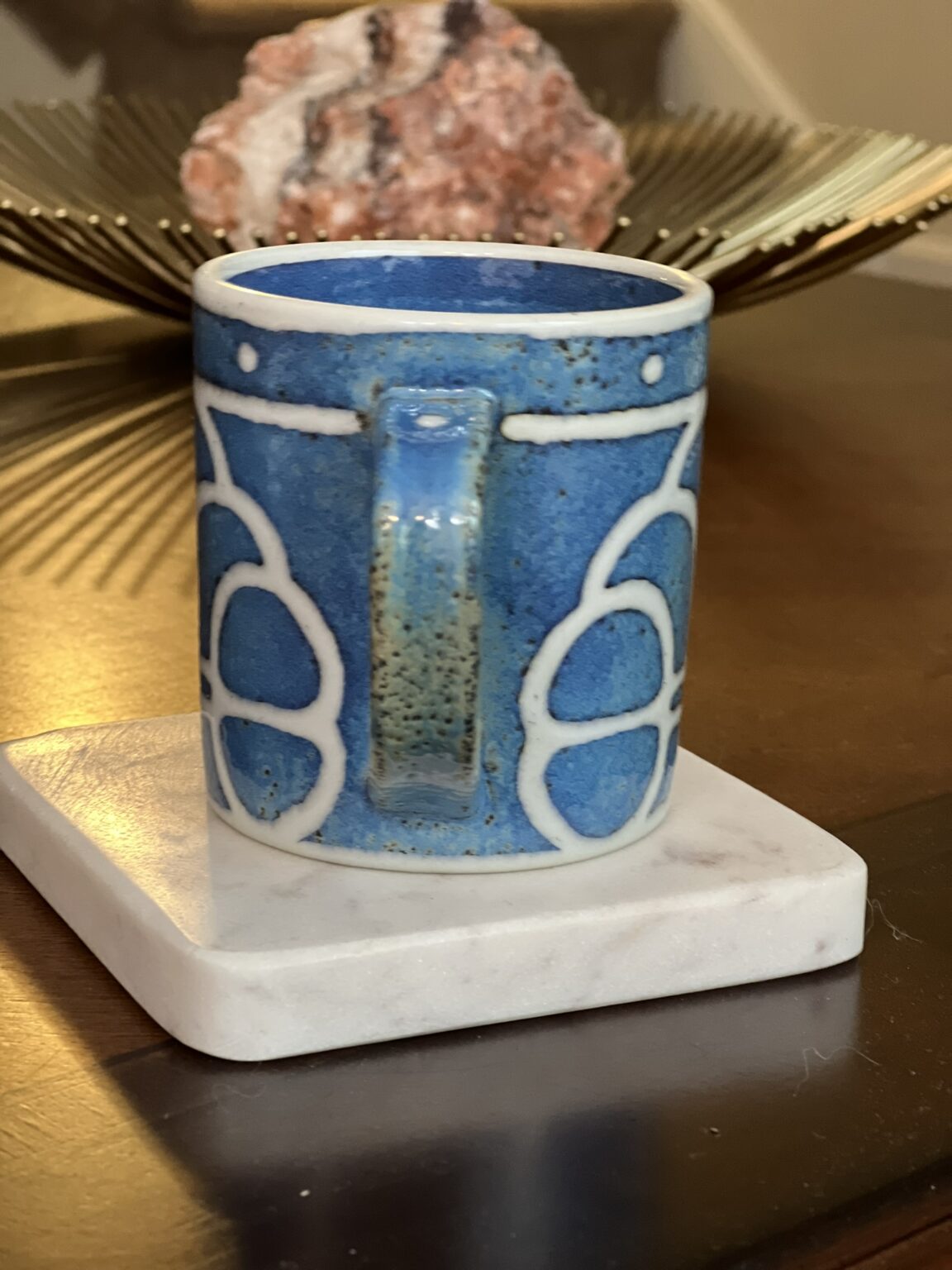 A blue ceramic mug with white geometric patterns sits on a white marble coaster atop a wooden table. In the background, a decorative tray with a large pinkish rock is visible.