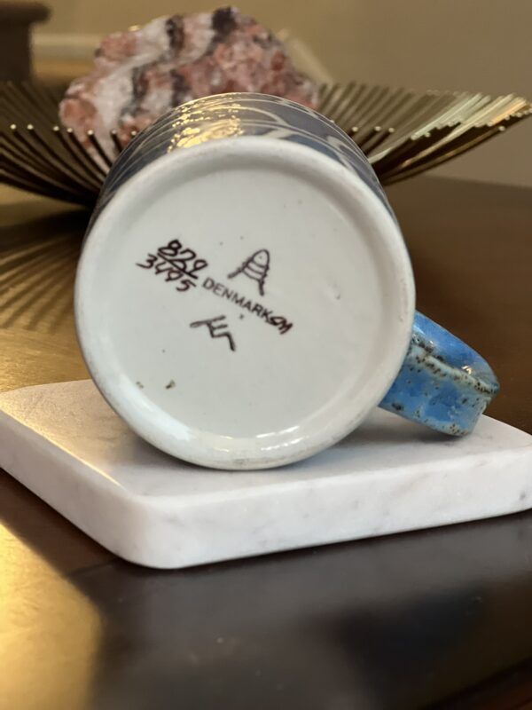 A blue ceramic mug is shown upside down, revealing a makers mark with the word DENMARK and a beehive symbol. The mug rests on a white square coaster, with a decorative object in the background.