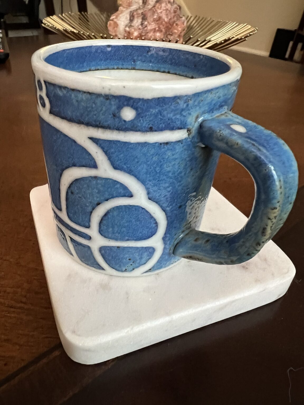 A blue ceramic mug with white abstract patterns sits on a square white coaster on a wooden table. In the background, there is a decorative object and part of a dining area visible.