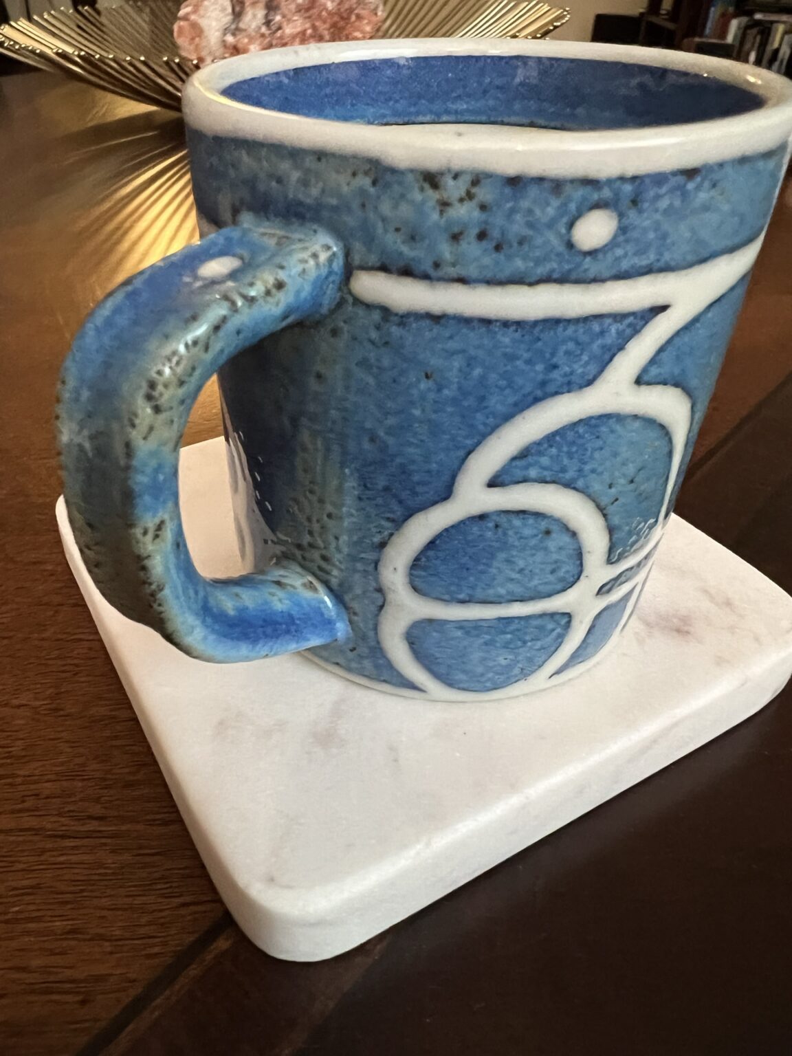 A blue ceramic mug with white abstract patterns sits on a white square coaster, placed on a wooden table. A decorative bowl and bookshelf are visible in the blurred background.