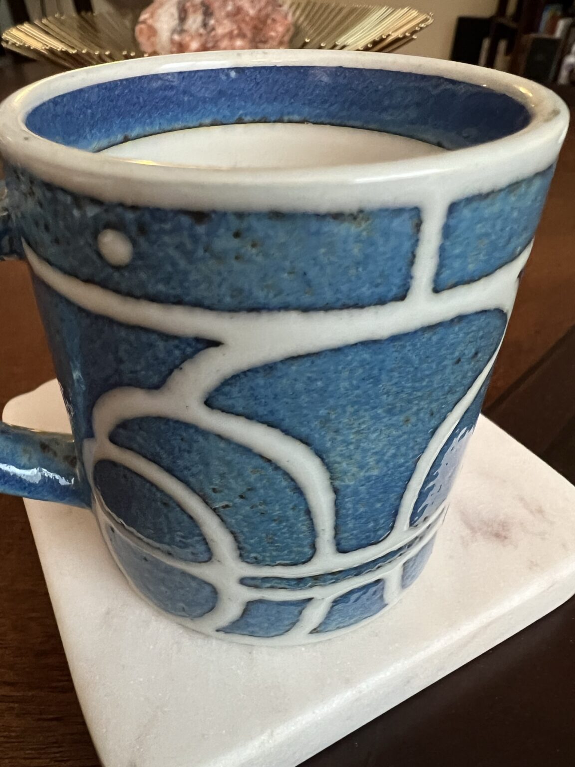 A close-up of a blue ceramic mug with white geometric patterns, filled with a creamy beverage, sitting on a white coaster on a wooden table. Blurred objects are visible in the background.