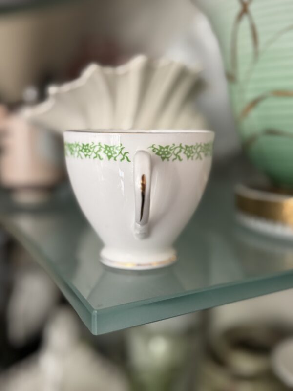 The English Ivy Teacup, crafted from white porcelain and accented with a green vine motif on the rim, rests on a glass shelf with its handle forward. A brown stain lies beneath, while blurred ceramics and pastel décor color the background.