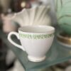 An English Ivy Teacup, featuring a green floral pattern around the rim, rests on a glass shelf with decorative items—including a large light green accent and a shell-shaped white piece—blurred in the background.