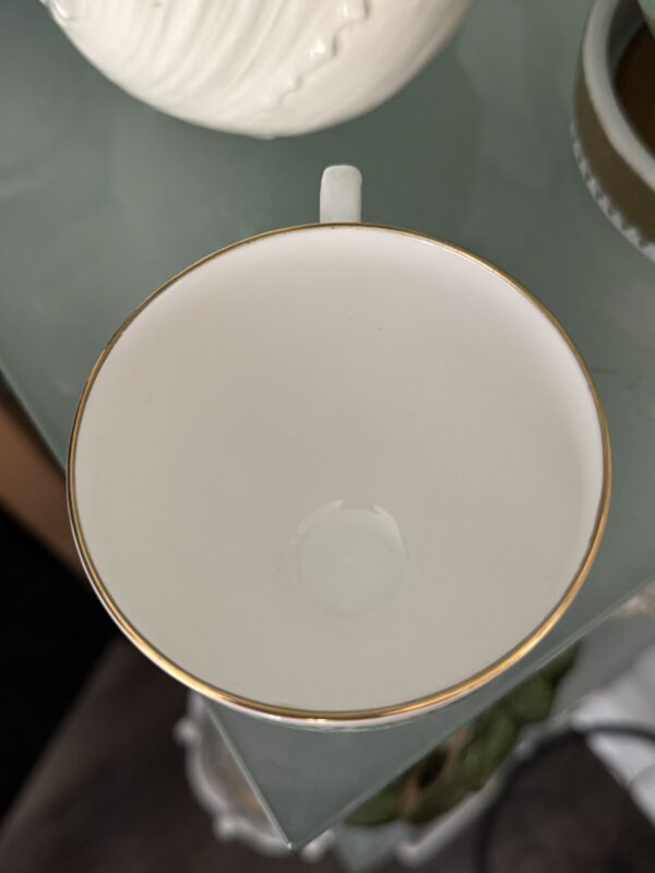 The English Ivy Teacup, a white porcelain cup with a thin gold rim, rests empty on glass. Its handle is visible at the top, and both a white ceramic object and English Ivy leaves appear softly in the background.